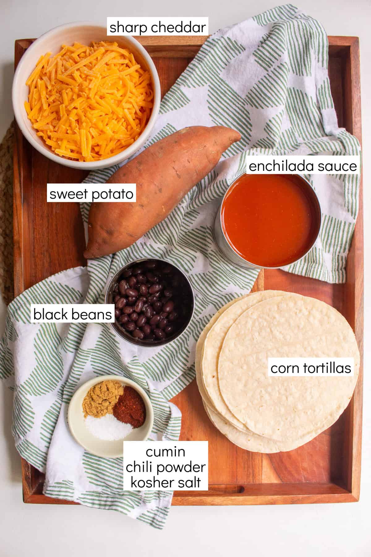 Ingredients for the recipe are displayed and labeled with white text boxes including the words sharp cheddar, sweet potato, black beans, enchilada sauce, cumin, corn tortillas, chili powder, and kosher salt.